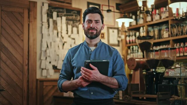 Caucasian bearded barman in black apron with digital tablet in hands smiling positively while posing at favorite workplace. Blurred background of bar counter with alcohol bottles arranged on shelves.