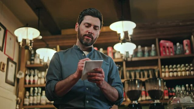 Positive bearded restaurant worker using pen and notepad while engaging in friendly conversation with clients. Attractive man in blue uniform writing order and wishes of customers during visit.