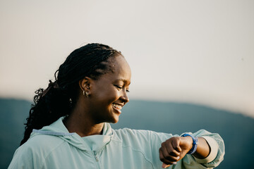 Confident African American woman running outdoors, syncing her pace with a smartwatch for optimal...
