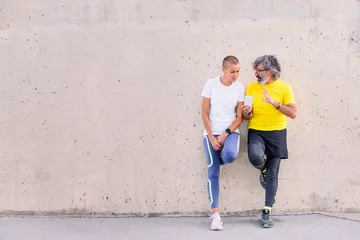 Foto op Plexiglas senior sports man and young sports woman talking and using mobile phone leaning an a wall, concept of technology of communication, copy space for text © Raul Mellado