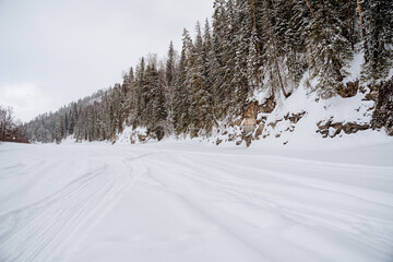 Winter road along the bed of a frozen river, cold weather, winter forest, taiga. White snow, snowdrifts on the river, mountainous terrain.