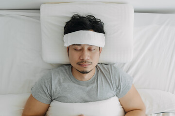 Top view of Asian man got sick with towel on forehead, man flu, man symptoms, resting alone and sleeping lying on white bed in apartment.
