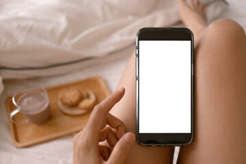Top view of phone showing white screen display, woman using and putting mobile on her legs, sitting on white bed white eating cookie and coffee.