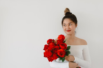 Asian Thai Korean woman wear red lips holding red roses, Happy smiling looking at empty space, standing isolated over white background wall with flowers in hand on Valentines day.