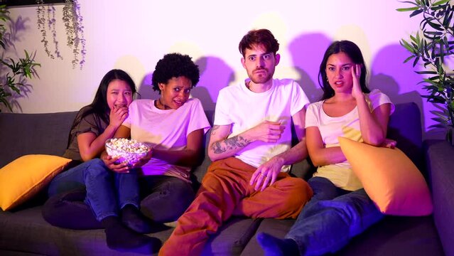 Multi-ethnic friends on a terror movie night at home sitting on the sofa eating popcorn and covering faces
