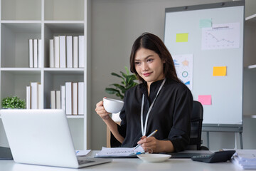 Beautiful smiling Asian businesswoman working using laptop and drinking coffee Asian businesswoman working with finance and calculator in her office