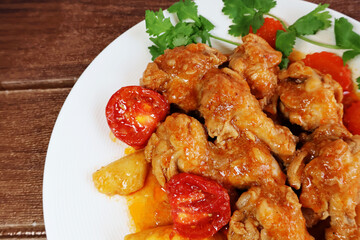 Closeup of stewed chicken legs with tomato sauce, pieces of pineapple, carrots, tomatoes in a cream colored porcelain plate on a dark brown wooden surface.

