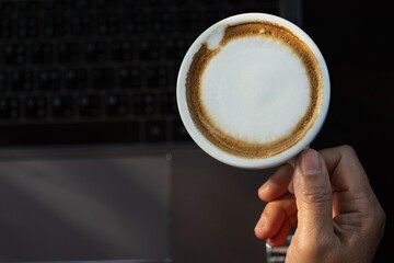 Business women hand holding white latte coffee cup and laptop on black table background.Hot coffee cup with laptop on desk when work from home.