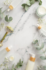 Natural body care ensemble. Top view vertical shot featuring gua sha roller, skincare essentials, cream and serum bottles, eucalyptus, and gypsophila bloom on a marble background with space for text