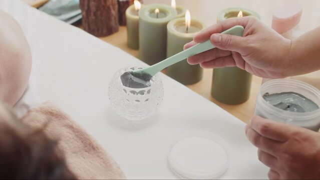 Close up of hands of unrecognizable woman preparing gray cosmetic clay for applying on face of male client