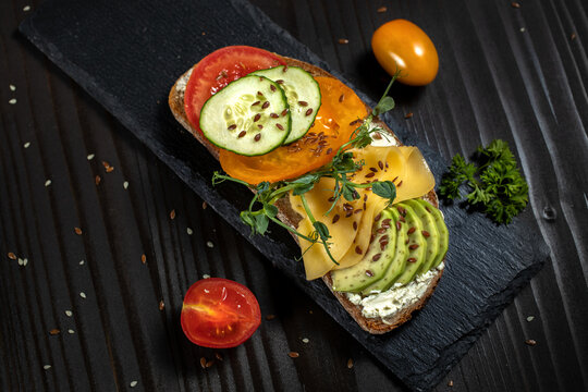 Open vegetarian sandwich with avocado, cheese, cucumber and cherry tomato on a black background