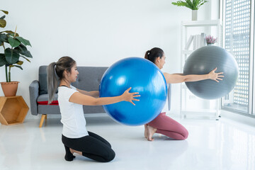 Fototapeta premium Teacher assists to do yoga pose training stretching body workout on mat with fitness ball at home for good health and body shape. Two asian fitness woman practicing yoga lesson course.