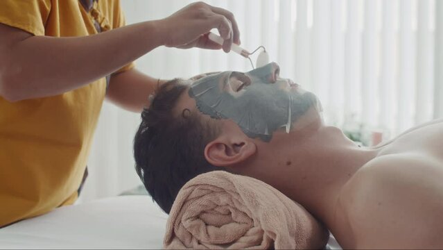 Side view of young man relaxing on couch while specialist doing beauty procedure on his face with jade roller and sheet mask