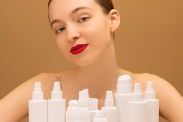Obraz na płótnie Canvas Close-up portrait delighted young female with red lips poses sitting at a table with cosmetic bottles and sprays on isolated peach background. The concept of a daily beauty routine. Beauty ad