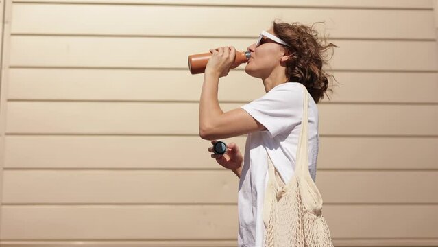 Cheerful millennial young woman opens a reusable metal bottle and drinks fresh water on sunny summer day outdoors on modern building wall background. Zero waste, eco-friendly, no plastic concept.
