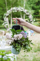 Female hand holds a bucket with lilac flowers, purple giant onions, and other flowersin front of a wedding arch. Bouquet of flowers as adecorative element. Vertical orientation - 706327726