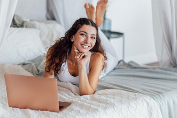 An attractive young woman in a white fitness suit is lying on the bed and typing on a laptop in the...