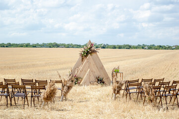 A rustic-style wedding ceremony venue nestled in a rural setting. A triangular wooden arch, adorned with flowers, graces the golden, mown field