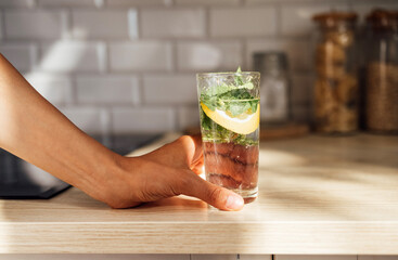 Close-up of female hand holds a refreshing lemon mojito on a wooden table on a kitchen at the home.