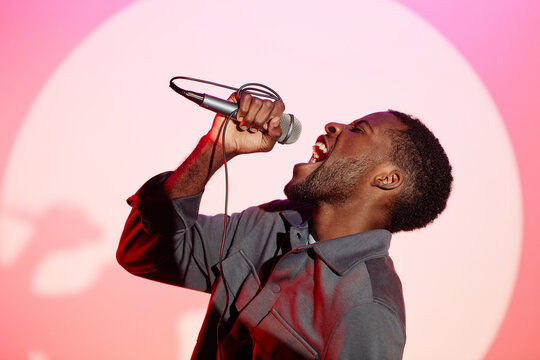 Side view of African American man standing at pastel pink wall of karaoke club and singing passionately into microphone