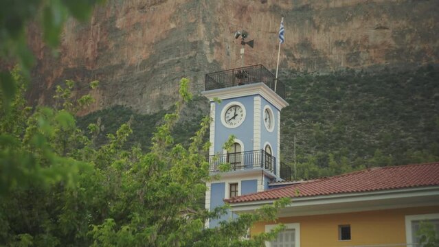 Vibrant blue clock tower with Greek flag below tall cliffs of Leonidio Greece