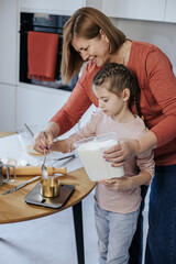 A Caucasian woman of 60 years old with a girl of 8 years old is pouring flour for dough in a light kitchen, there are various kitchen utensils on the table