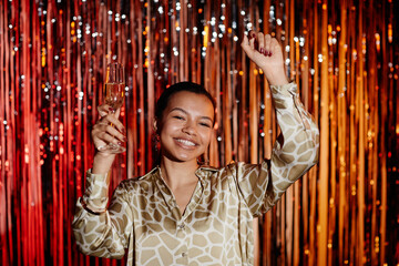 Medium shot of joyful Black woman in stylish shirt at nightclub with hands in air holding champagne...
