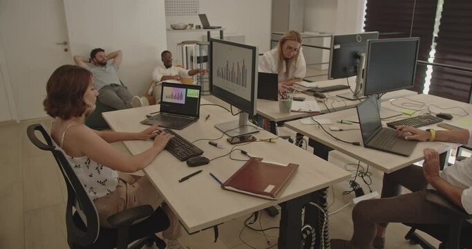 Three people working on the computers while two other coworkers are resting on the cozy office sofa, making a break from work