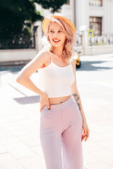 Young beautiful smiling blond hipster woman in trendy summer clothes. Carefree female posing on the street background at sunny day. Positive model outdoors at sunset. Cheerful and happy in hat