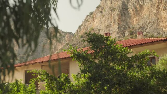 Wind blows green tree and shrub as sunlight illuminates house below towering cliffs of Leonidio Greece