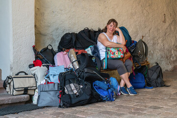 A smiling middle-aged woman is sitting on a bench, next to a huge amount of luggage, many bags,...