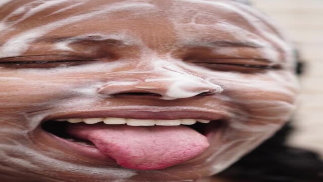 Vertical close-up of a cheerful young black woman making funny faces with soap suds on her face in the shower at home. Latin female having fun in her skin and body care early morning routine.