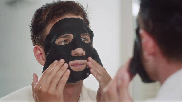 Over the shoulder view of young man applying black charcoal mask on face in front of mirror during morning skin care procedure