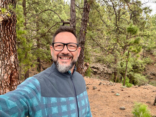 One adult man taking selfie picture with forest park woods in background. Mature using mobile phone to take picture during weekend activity wearing glasses and enjoying outdoor leisure activity travel