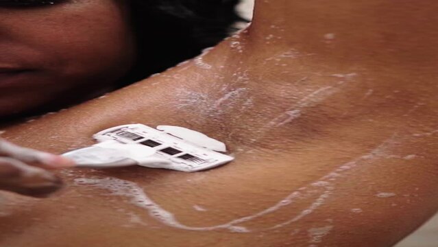 Young black woman shaving her armpits in the shower. Latin female removing her hair with a soft razor in the bathroom of her home. Body care routine and skin cosmetics. Vertical close up.
