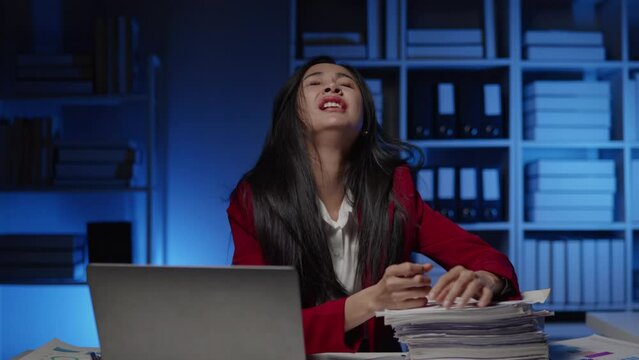 Businesswoman in red suit is seriously stressed working overtime, bored and burnout due to office syndrome on laptop and paperwork in the office.