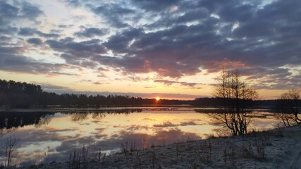 Fototapeta na wymiar The sun rises from behind the forest on the far side of the lake and brightly colors the cloudy sky. Trees and clouds are reflected in the water. The grass on the shore is covered with frost