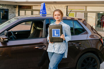 Young female learner driver hold clipboard with L sign standing next to brown vehicle with blue L...