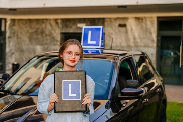 Young charming female driver posing with L Plate on the roof of Vehicle . Driving school and people concept. Young beautiful happy woman holding folder with L sign near training car.