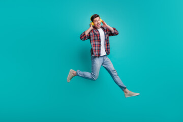 Full length photo of cool funky man dressed plaid shirt jumping high enjoying music headphones isolated turquoise color background