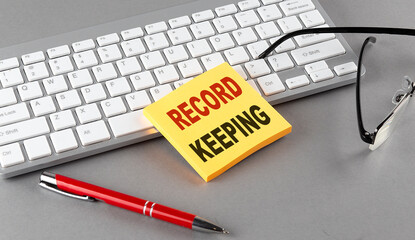 RECORD KEEPING text on a sticky with keyboard, pen glasses on grey background