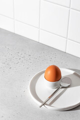 Boiled egg in an egg stand and spoon on white tile kitchen background with text space. Light...