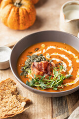 Pumpkin cream soup decorated with cream, pumpkin seeds, bacon, microgreens and olive oil on brown textured background with ingredients