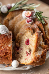 Christmas bundt cake decorated with glaze, cranberries and rosemary on beige table cutted close up. Homemade cake recipe