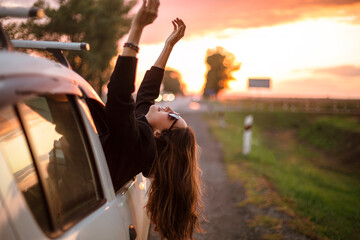 Young carefree woman stretching out of car window while driving through countryside at sunset