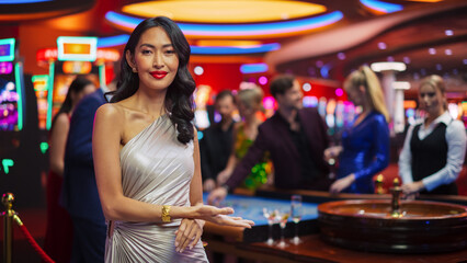 In a Modern Casino Hall: Gorgeous Asian Brunette Woman Posing Confidently, Looking at Camera, Pointing at a Placeholder Space for Advertising or Commercial Materials for a Gambling Industry