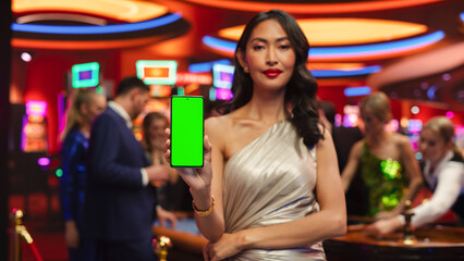 Modern Casino Setting: Gorgeous Asian Brunette Woman Posing with a Mobile Phone with a Green Screen...