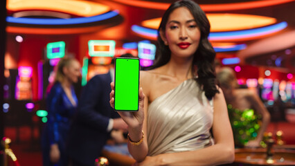 Advertising Template with an Asian Female Showing a Smartphone Device with a Green Screen Mock Up Display with Trackers. Professional Beautiful Model in a Casino with People Gambling in the Background