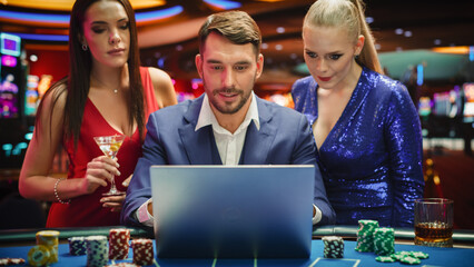 Portrait of a Happy Man with Two Female Partners Betting Online on a Laptop Computer while Sitting in a Luxury Casino. Lucky Man and Glamorous Women Celebrating Blackjack Winning Jackpot . 