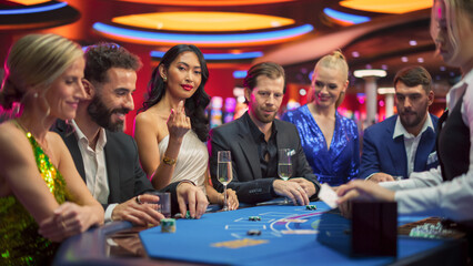 Portrait of a Glamorous Asian Woman at a Blackjack Gambling Table in a Modern Casino. Glamorous Successful Lady Looks at the Camera Calls to Join her at the Table, Smiling while People Win. - obrazy, fototapety, plakaty